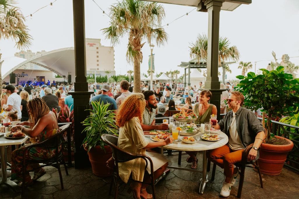 A lively outdoor dining scene featuring a group of friends enjoying meals and drinks at a table, with a music event and audience in the background.
