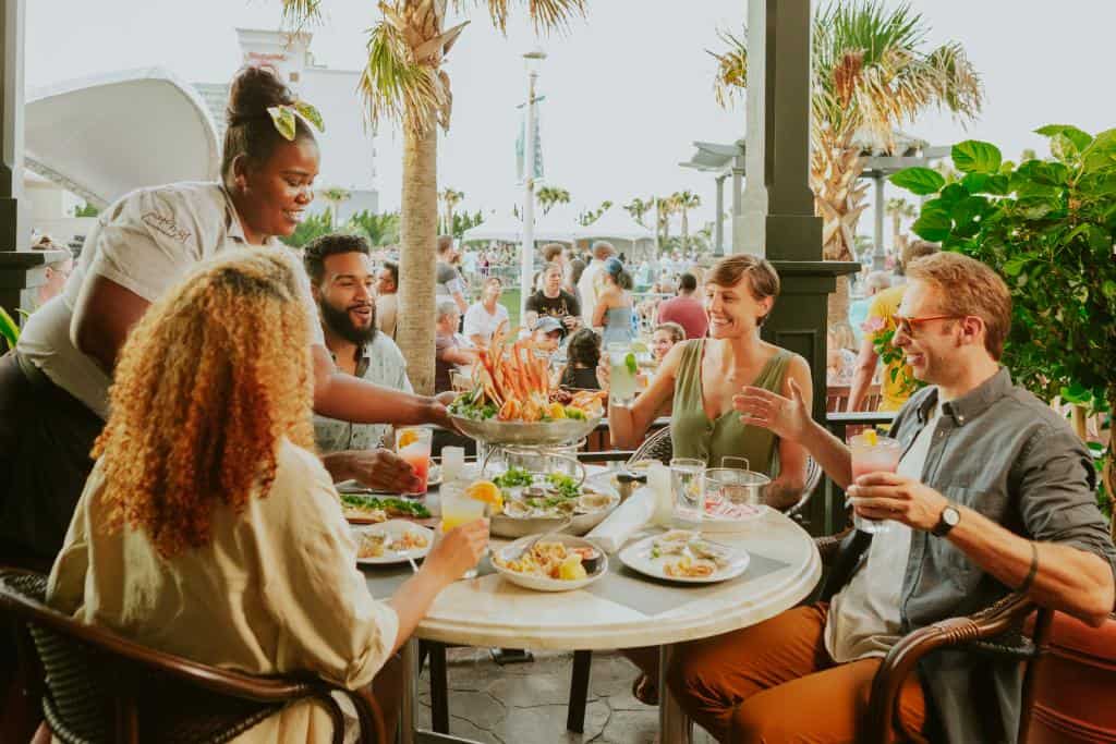 A group of five friends enjoys a vibrant outdoor meal at Catch 31 restaurant, featuring seafood and drinks, with a waitress serving them.