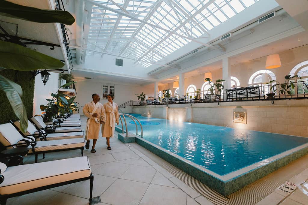 A couple in bathrobes walk beside a serene indoor pool in a well-lit spa, surrounded by plants and elegant seating.