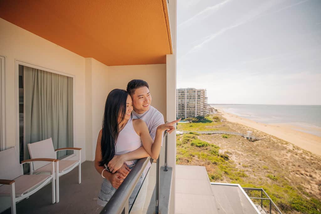 A couple enjoys a sunny day on a balcony overlooking a beach and ocean, with residential buildings in the background.
