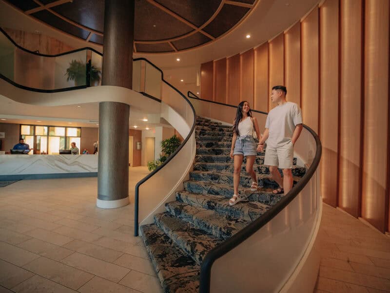 A couple walks hand-in-hand down a stylish curved staircase in a modern hotel lobby with contemporary decor.