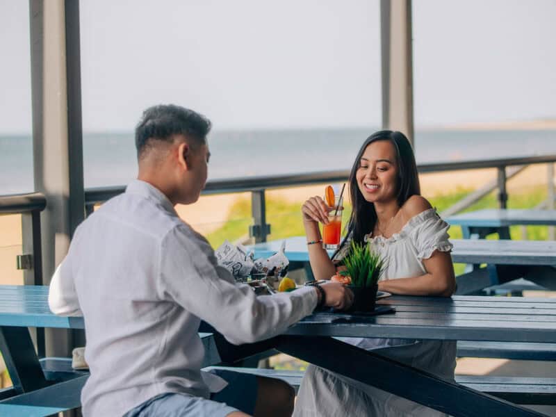 A couple enjoying drinks and food at a beachside outdoor cafe, with a scenic view of the ocean in the background.