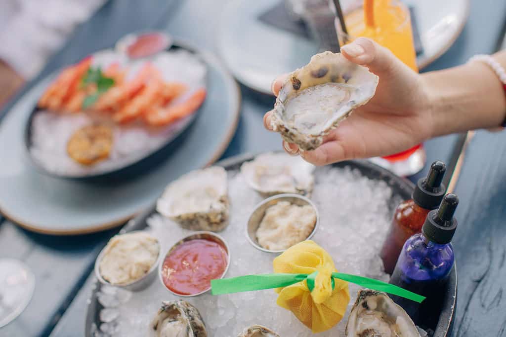 A hand holding a fresh oyster above a plate of oysters on ice, accompanied by sauces and a colorful garnish.
