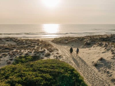 A couple walks along a sandy path towards the beach at sunset, with dunes and ocean waves in the background.