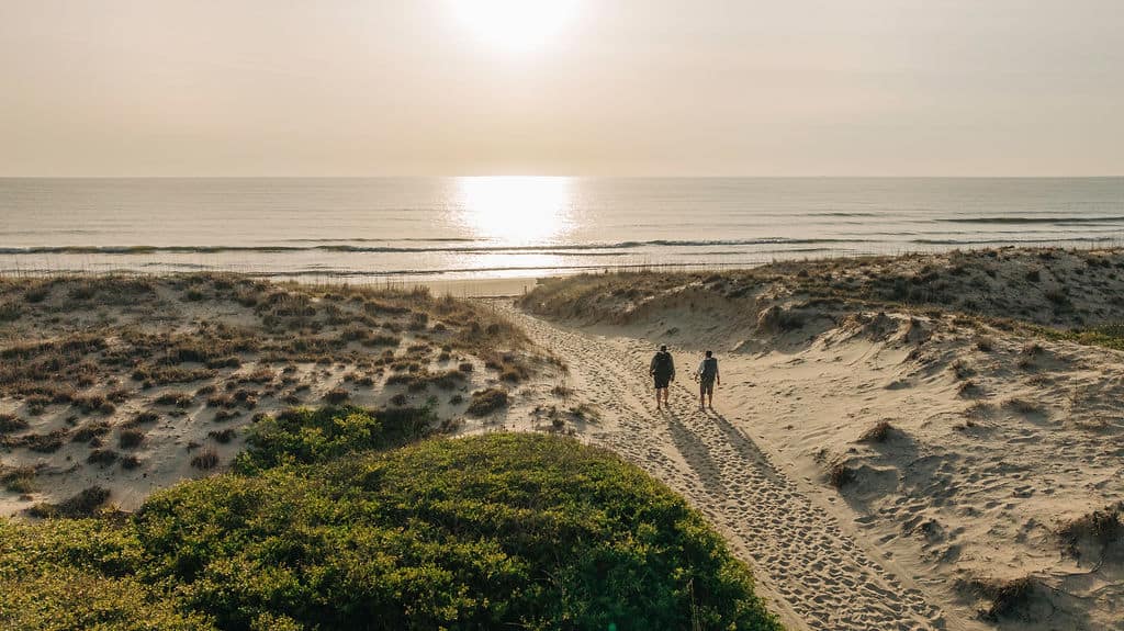 A couple walks along a sandy path towards the beach at sunset, with dunes and ocean waves in the background.