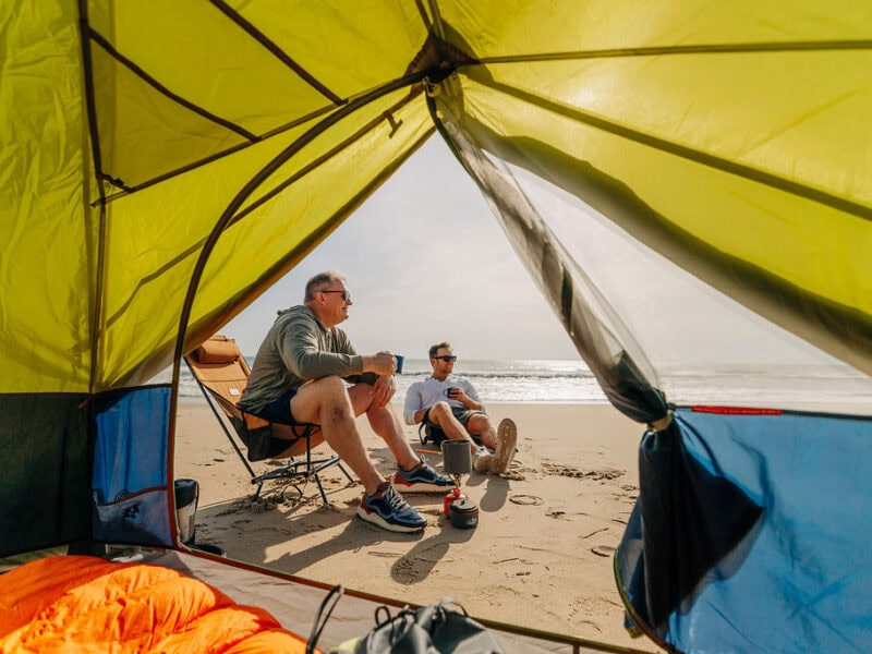Two men relaxing inside a beach tent, enjoying drinks and ocean views on a sandy shore.