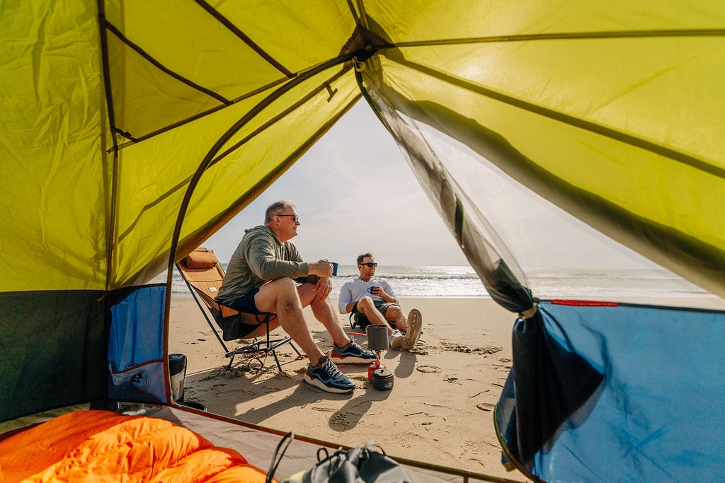 Two men relaxing inside a beach tent, enjoying drinks and ocean views on a sandy shore.