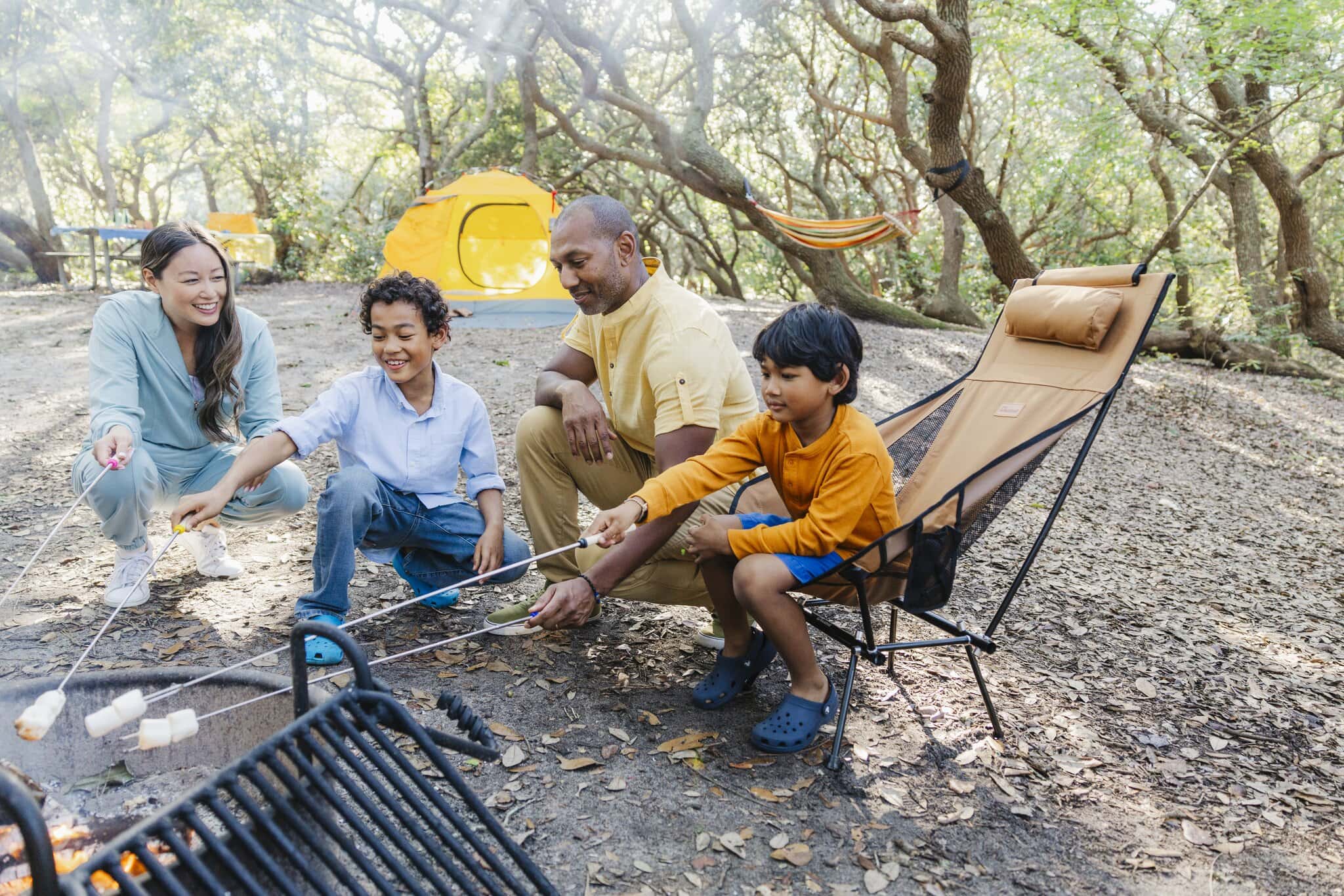 A family enjoys a camping trip, roasting marshmallows over a campfire in a wooded area with a tent and hammock nearby.
