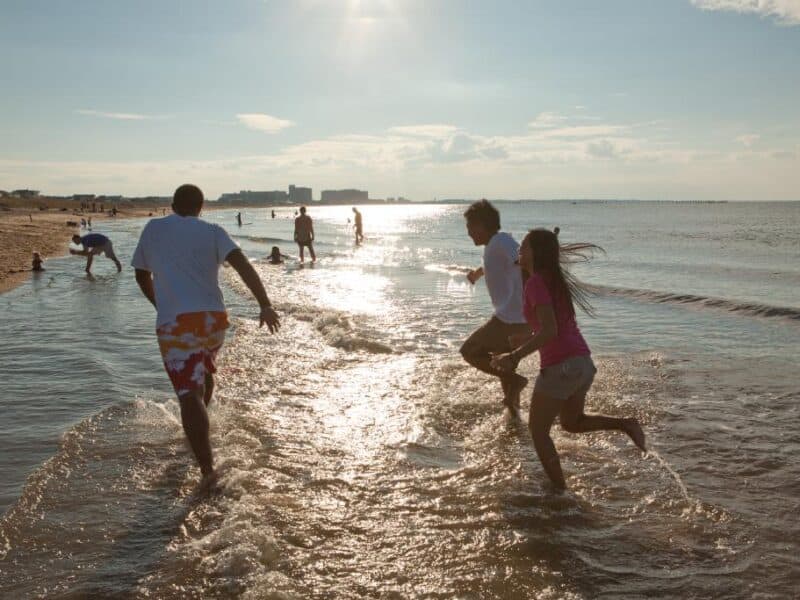 Three people play in the shallow water at First Landing State Park, enjoying a sunny day with gentle waves and distant beachgoers in the background.