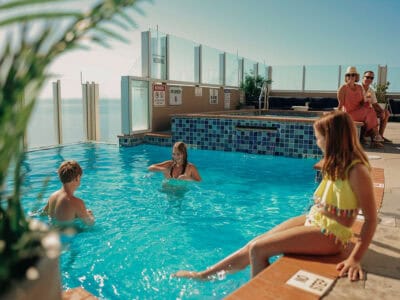 A family enjoys a sunny day at a rooftop pool, with children swimming and adults relaxing nearby, surrounded by scenic views.