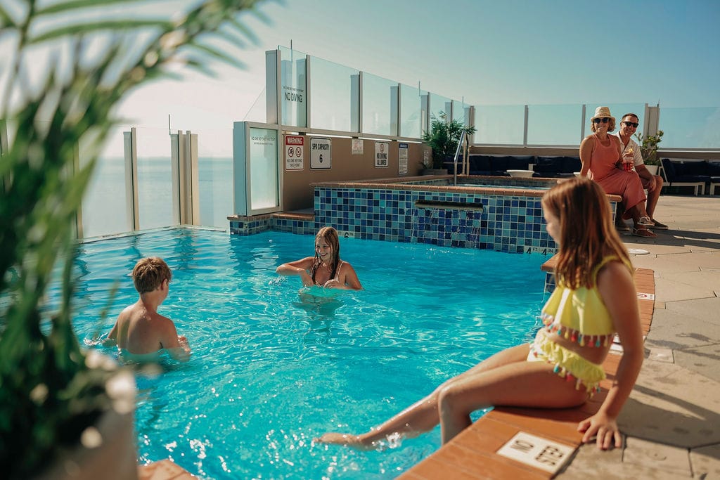 A family enjoys a sunny day at a rooftop pool, with children swimming and adults relaxing nearby, surrounded by scenic views.