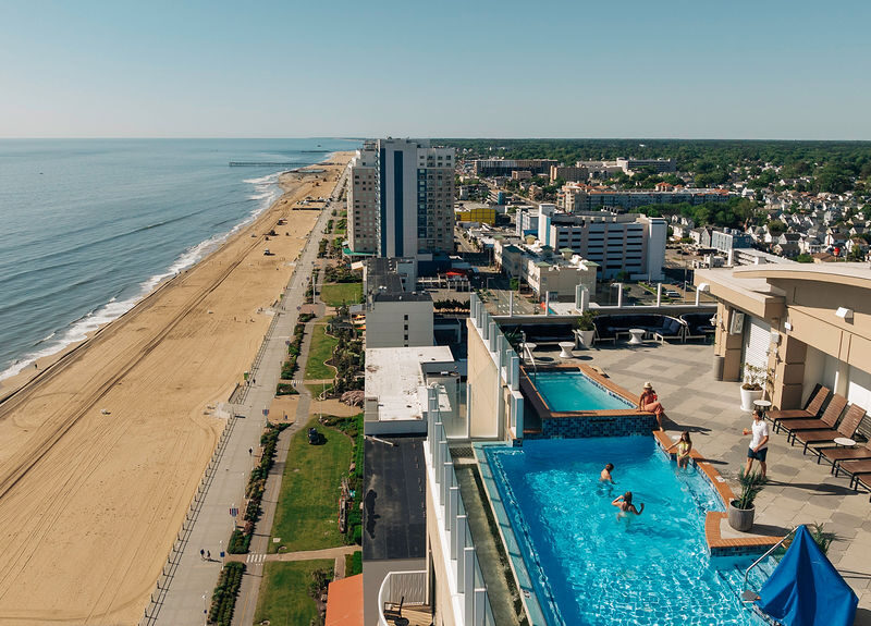 A panoramic view of a rooftop pool overlooking the beach and cityscape, showcasing a sunny day with people enjoying the water and sand.