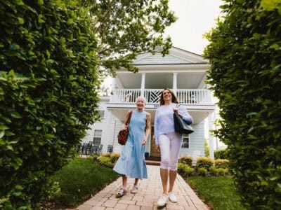 Two women walking down a path flanked by greenery, approaching a light blue house with a porch.