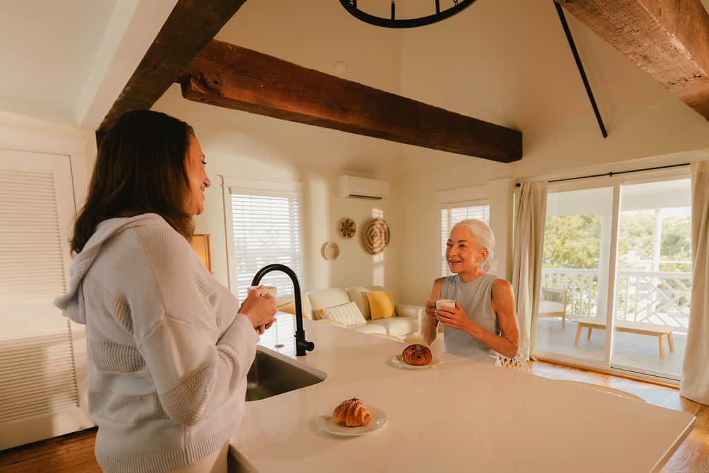 Two women engage in a cozy conversation in a bright kitchen, enjoying pastries and drinks, with sunlight streaming through large windows.