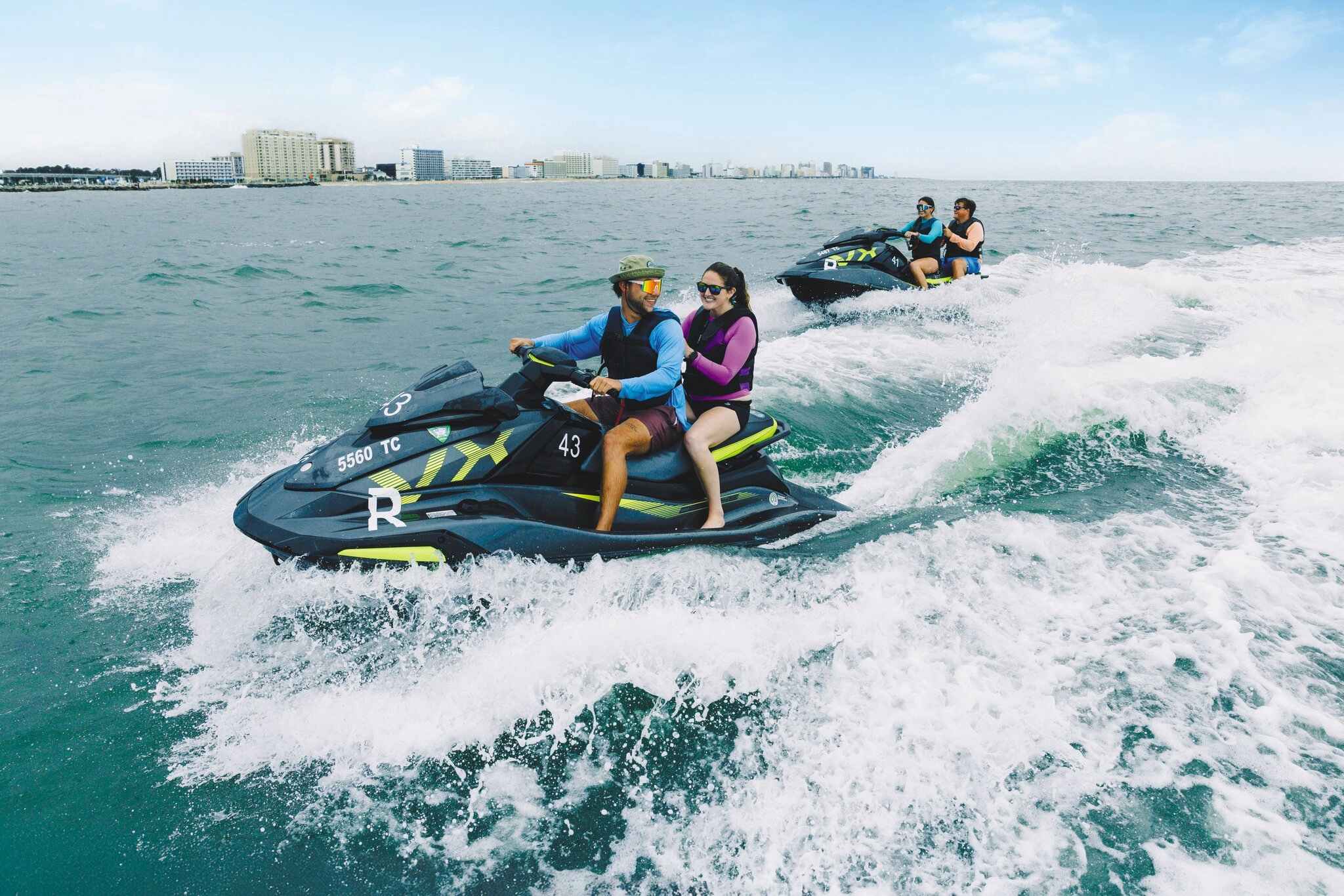Two people enjoy a thrilling ride on a jet ski, creating waves in the ocean, with a city skyline visible in the background.