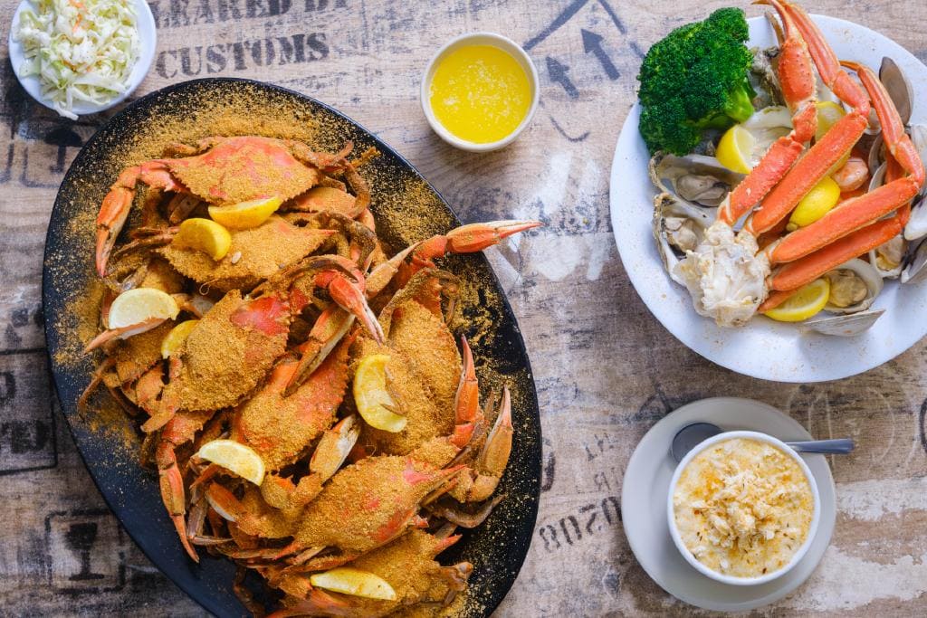A platter of seasoned crabs with lemon slices, accompanied by a side of broccoli, coleslaw, and a bowl of crab chowder.
