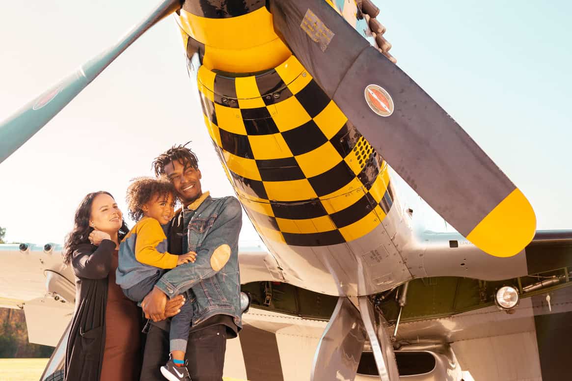 A family of three smiles together in front of a vintage airplane with a yellow and black checkered propeller.
