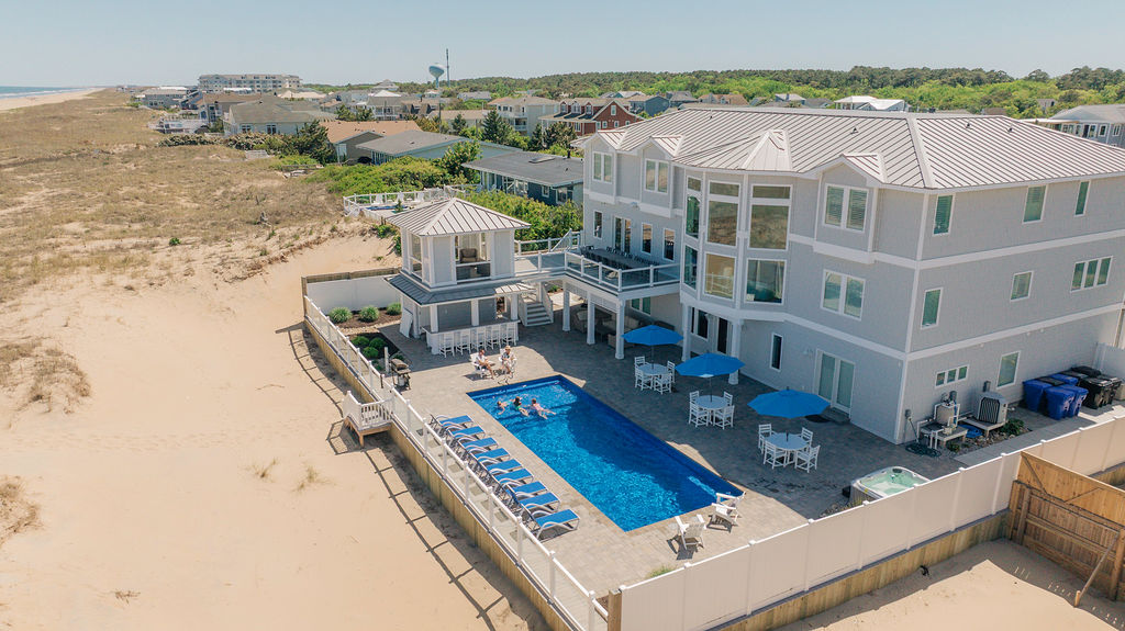 Aerial view of a beach house with a swimming pool, surrounded by lounge chairs and umbrellas, overlooking the sandy coastline.