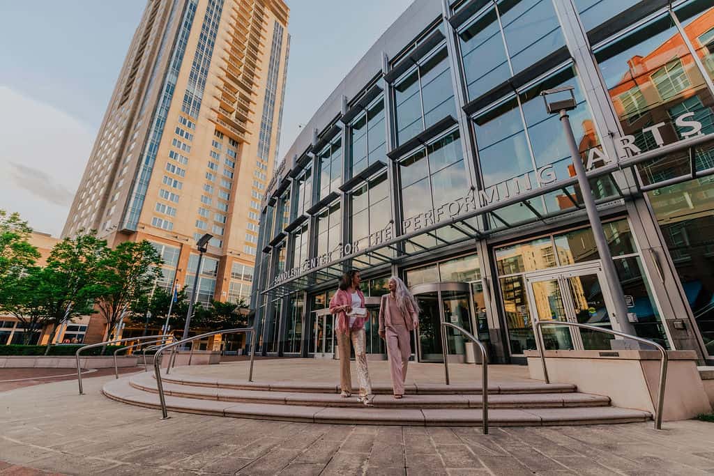 Two individuals walk up the stairs to the Sandler Center for the Performing Arts, surrounded by modern buildings and greenery.