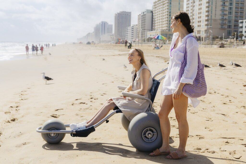 Two women enjoy a sunny day at the beach, one seated on a beach wheelchair with large wheels, while the other stands beside her holding a purple bag.