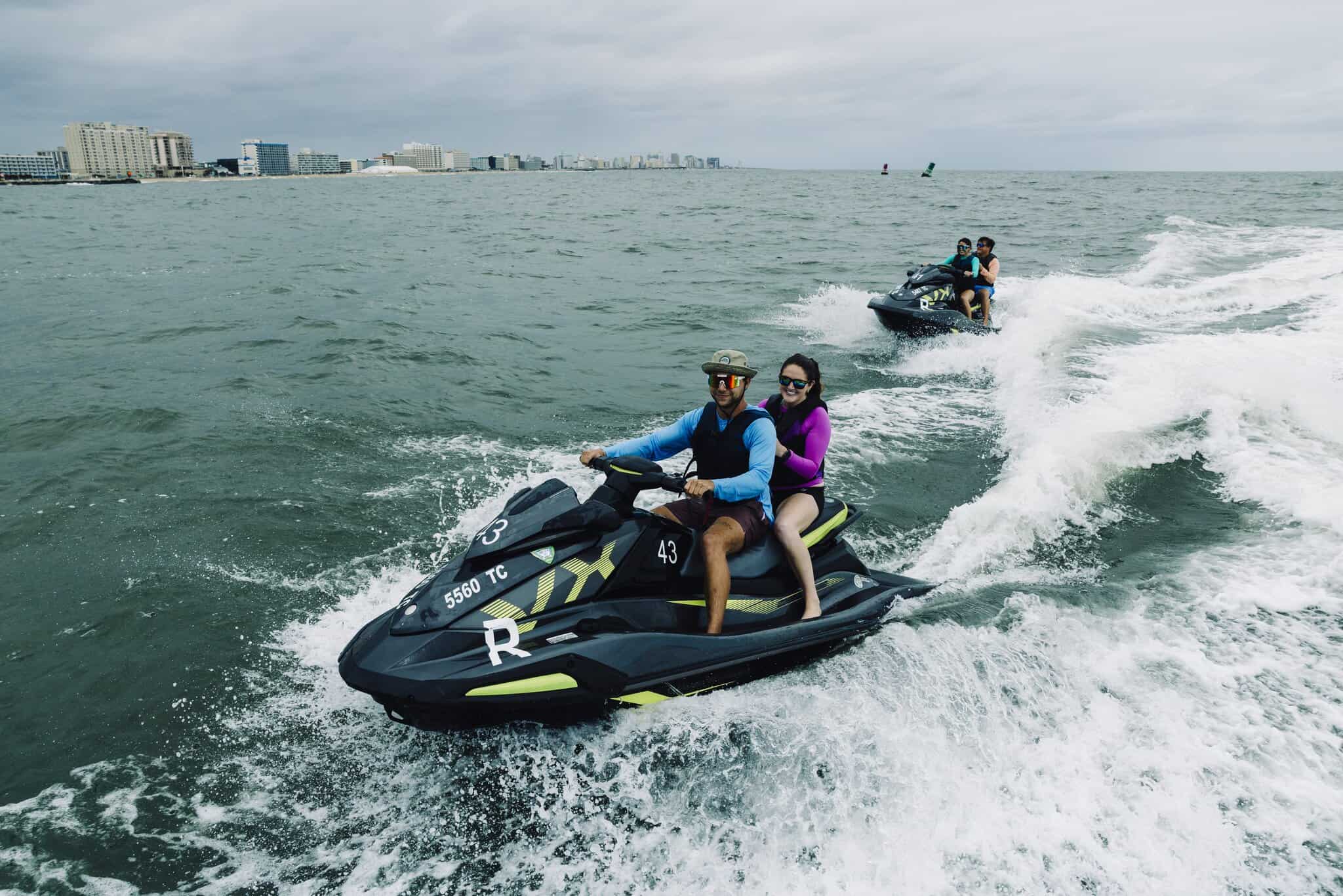 A couple enjoys jet skiing on the ocean, with city buildings visible in the background under a cloudy sky.
