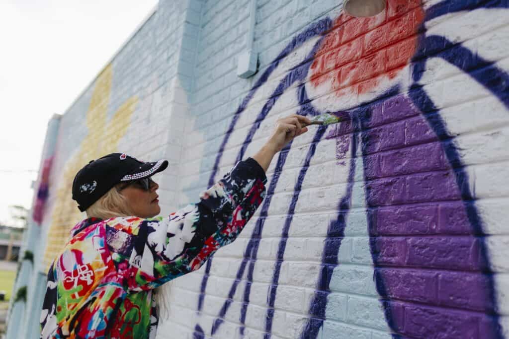 A woman with blonde hair paints a colorful mural on a brick wall, using a brush to add purple paint.