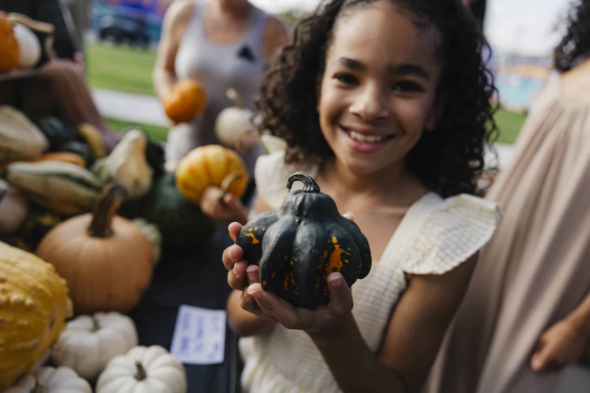 A cheerful girl holds a unique black and orange pumpkin at a fall market, surrounded by various other gourds.