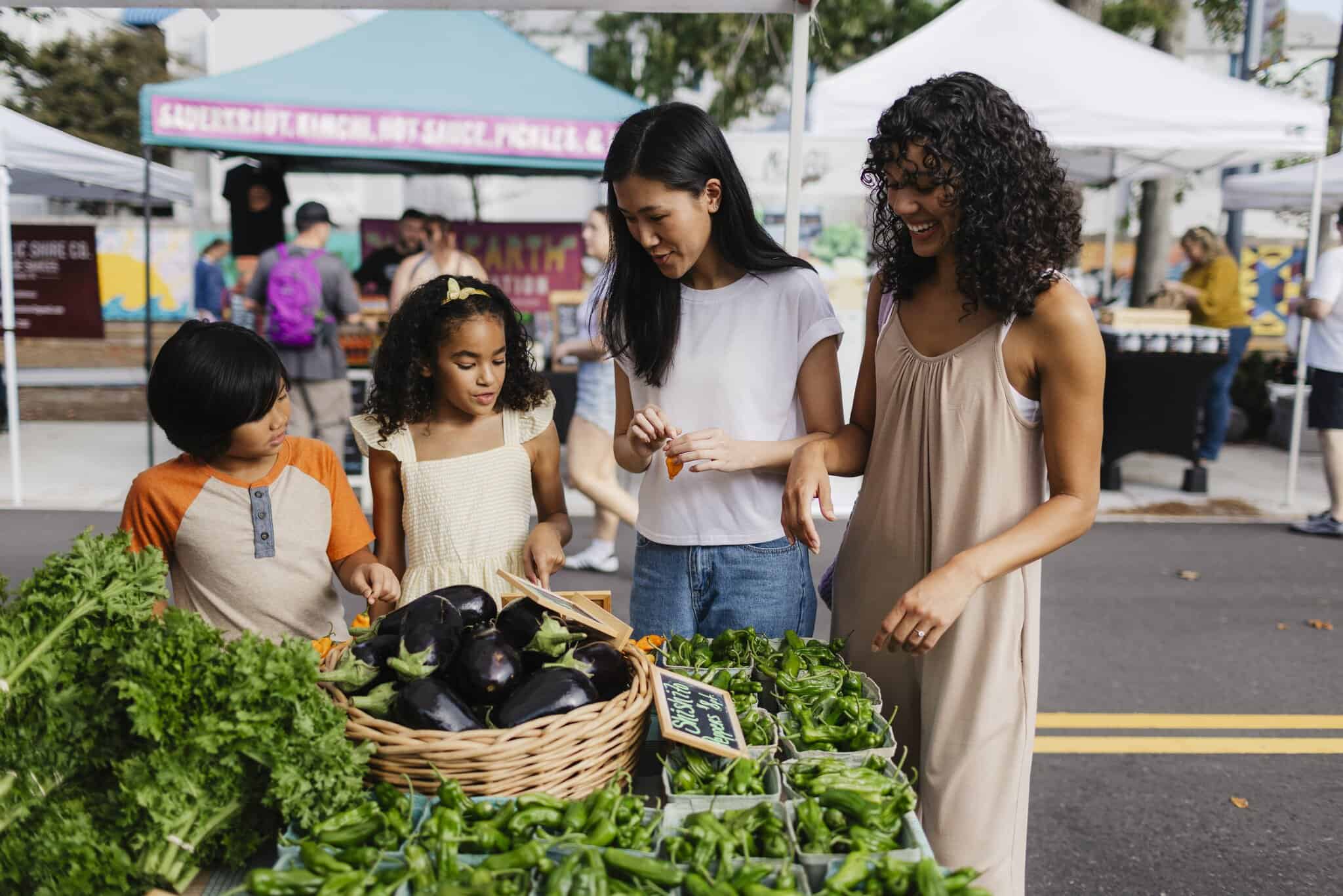 A family shopping at a farmers market, examining fresh vegetables like eggplants and peppers while enjoying the vibrant atmosphere.