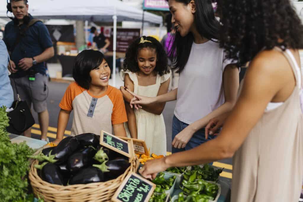 A group of children and adults joyfully interact while shopping for fresh vegetables at a vibrant farmers market.