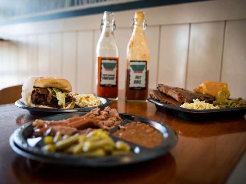 A table set with plates of barbecue meat, a pulled pork sandwich, and sides including coleslaw, green beans, and cornbread, accompanied by bottles of BBQ sauce.