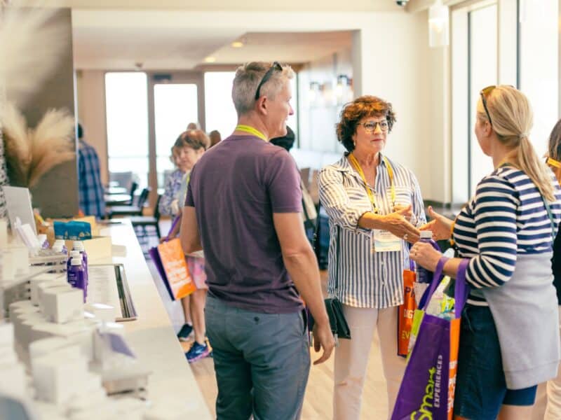 A group of attendees engage in conversation at a conference networking event, with colorful tote bags in a well-lit modern space.