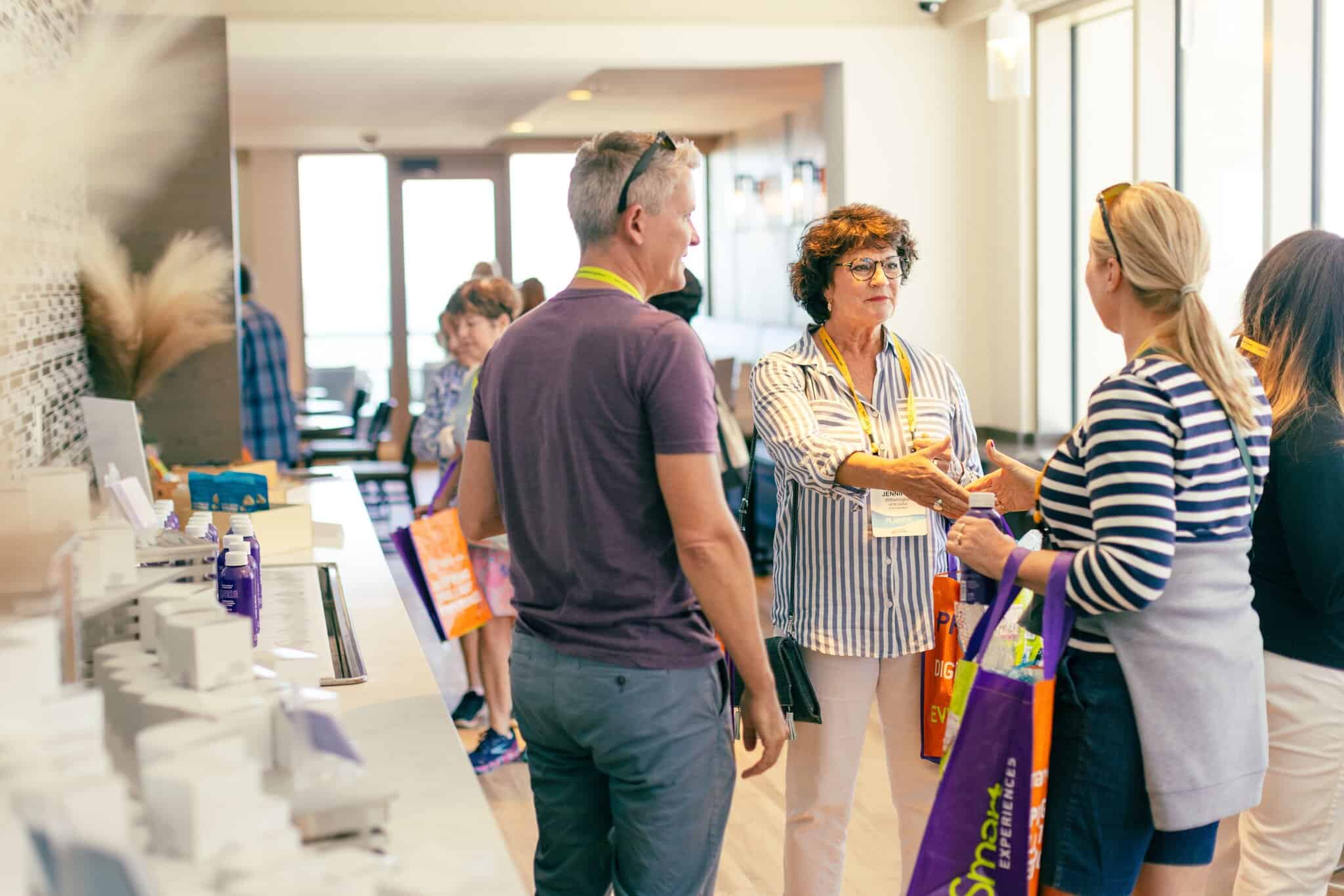 A group of attendees engage in conversation at a conference networking event, with colorful tote bags in a well-lit modern space.