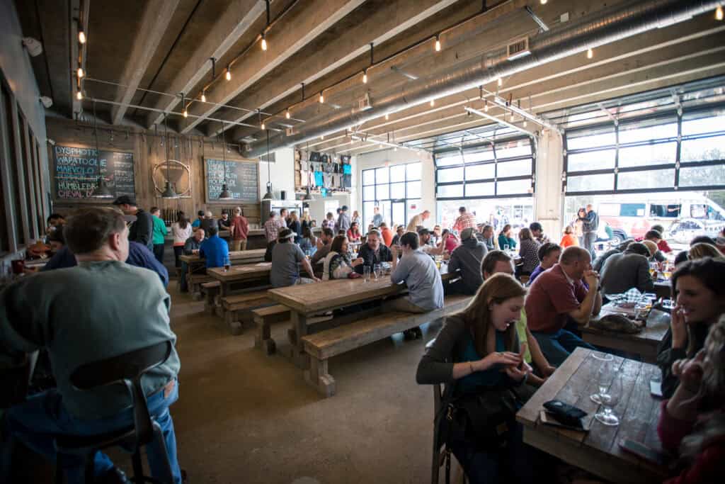 A lively brewery interior with wooden tables, patrons enjoying drinks, and a chalkboard menu on the wall, featuring a garage-style door.