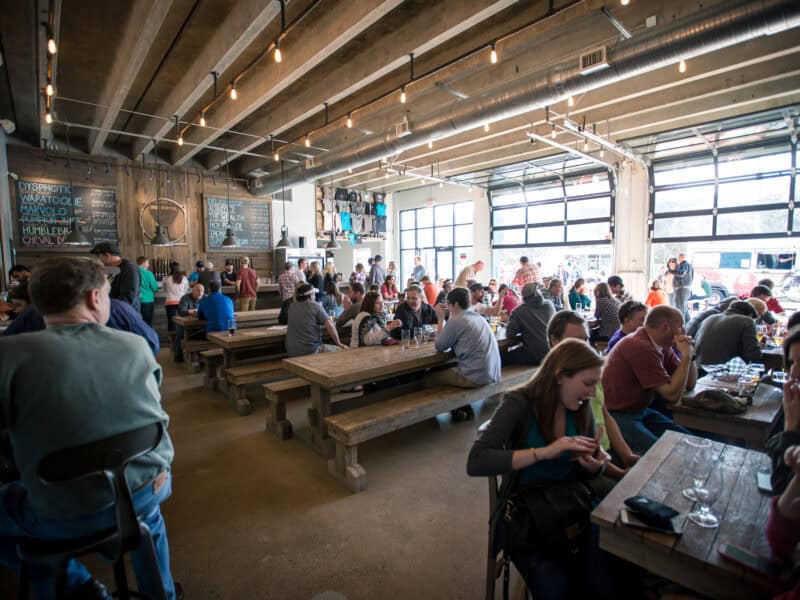 A lively brewery interior with wooden tables, patrons enjoying drinks, and a chalkboard menu on the wall, featuring a garage-style door.
