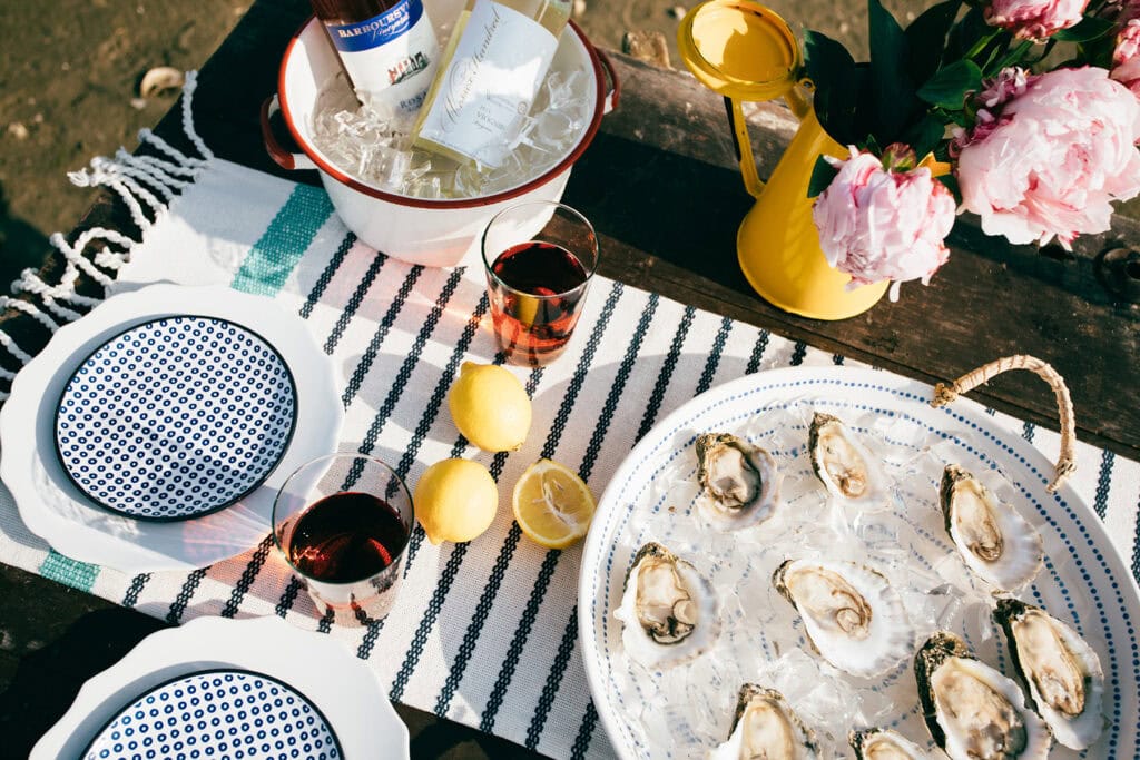 A beautifully arranged outdoor dining setup featuring oysters on ice, drinks, lemons, and floral decor on a striped tablecloth.
