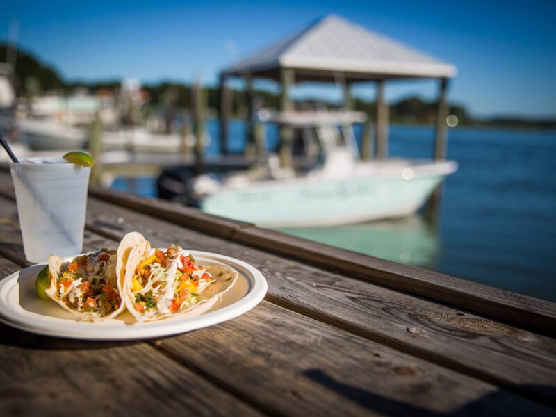 Fresh tacos with colorful toppings on a wooden table overlooking a marina, with a refreshing drink and boats in the background.