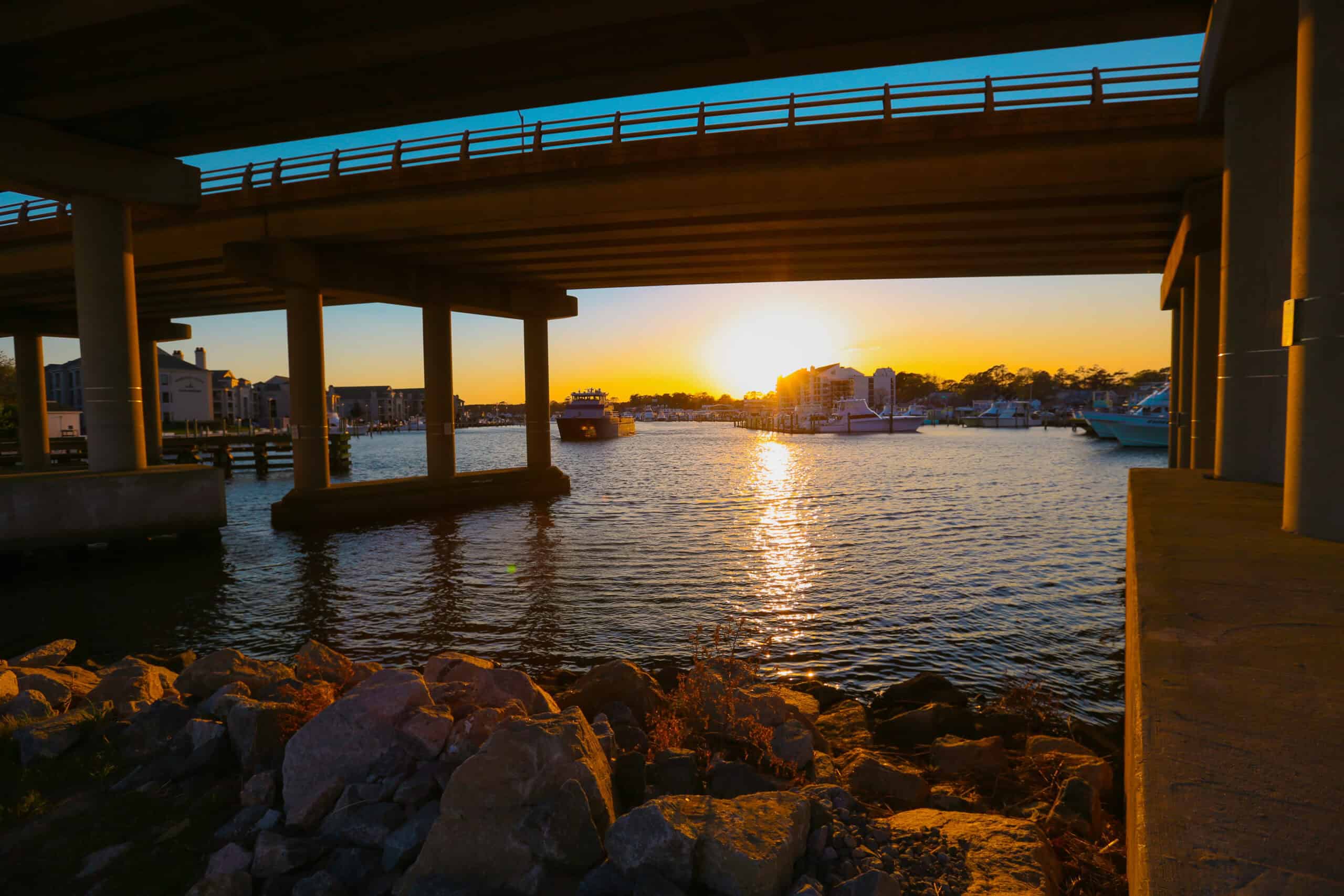 A sunset view under a bridge with boats in a marina, reflecting golden light on the water and stones in the foreground.
