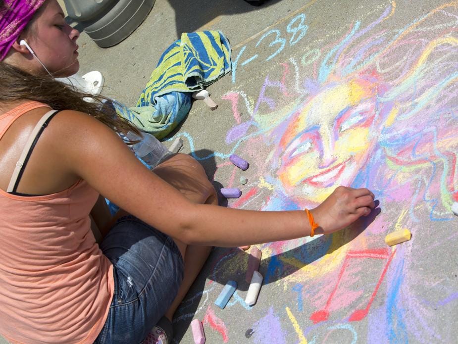 A young girl draws a colorful chalk portrait on pavement, surrounded by chalk pieces and a folded towel.