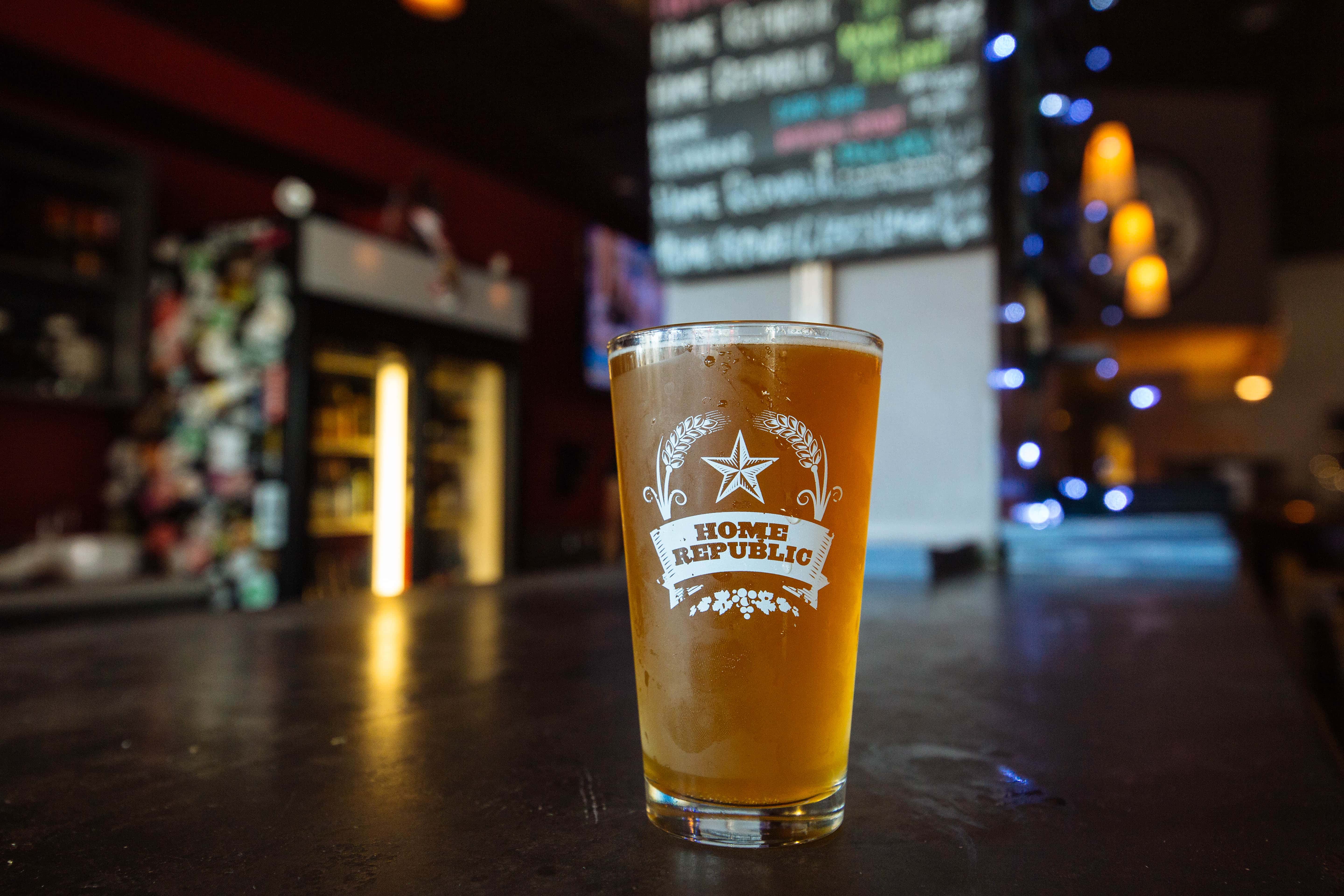 A glass of craft beer with a decorative logo sits on a bar counter, with ambient lighting and a menu board in the background.