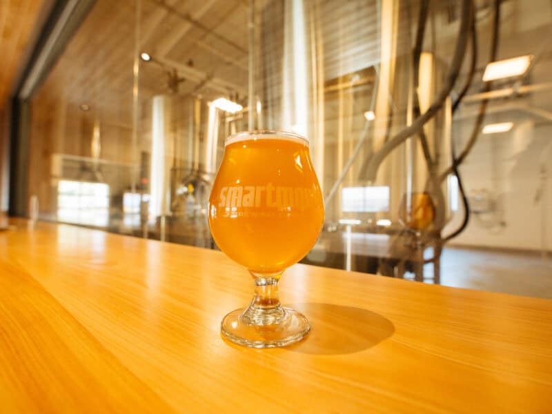 A glass of golden craft beer sits on a wooden bar, with brewing equipment visible in the background at Smartmouth Brewing Company.