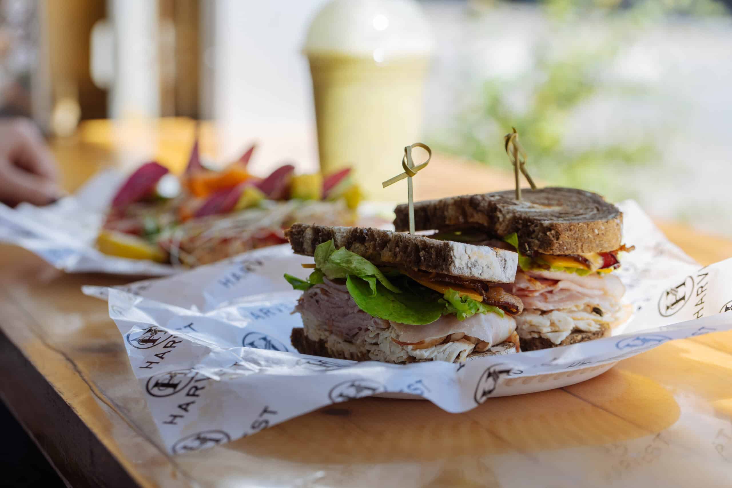 A close-up of a turkey and bacon sandwich on dark bread, garnished with lettuce and skewered, served on branded paper with a side of colorful vegetables.