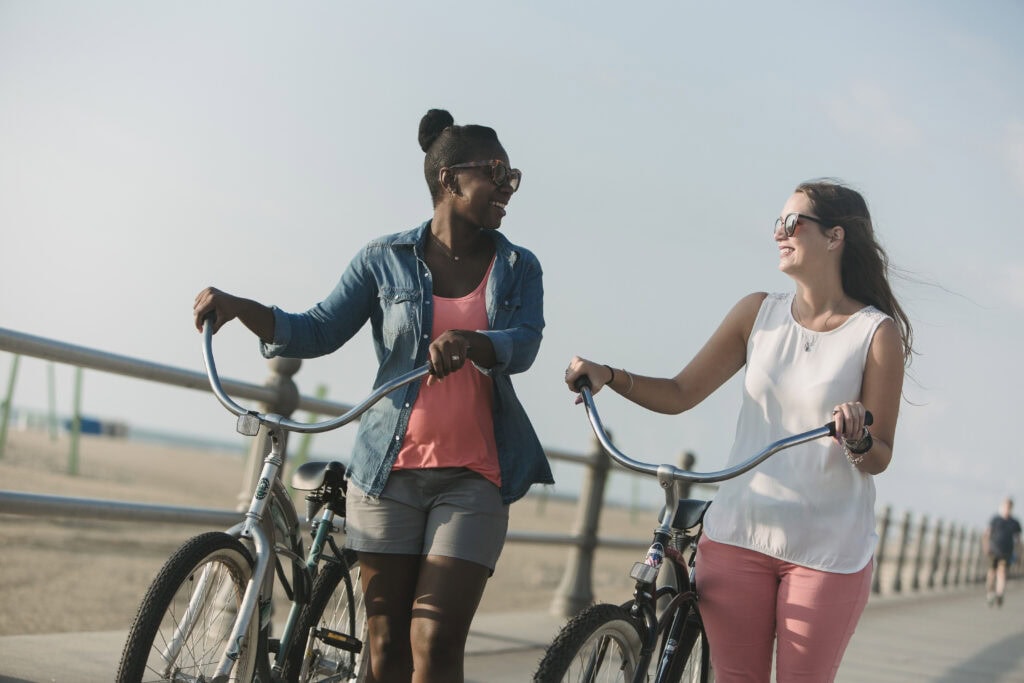 Two women joyfully walking with bicycles along a beachside promenade on a sunny day.
