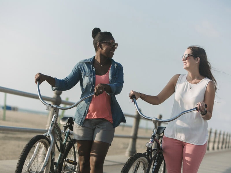 Two women joyfully walking with bicycles along a beachside promenade on a sunny day.