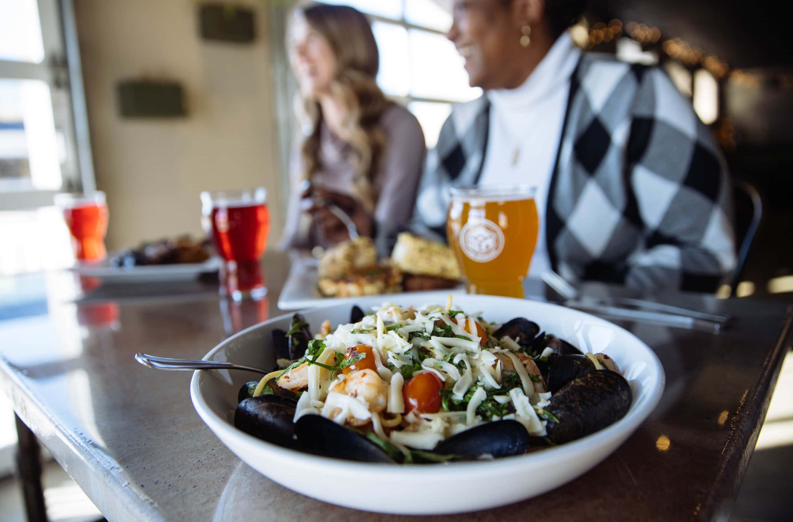 A bowl of mussels with shrimp, tomatoes, and cheese, surrounded by drinks and two women enjoying a meal at a restaurant.