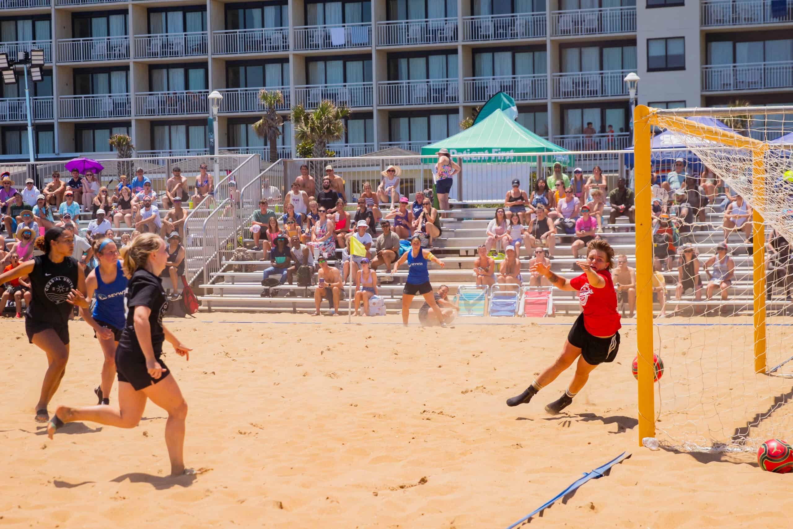 A sand soccer game in progress, featuring players chasing the ball near a goal, with a crowd watching from bleachers.