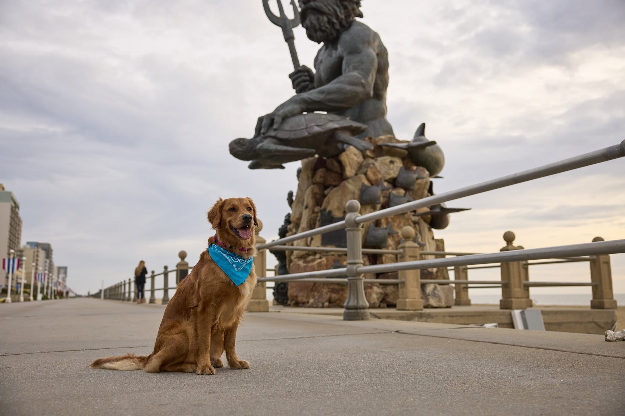 A golden retriever wearing a blue bandana poses on a boardwalk, with a large Neptune statue in the background.