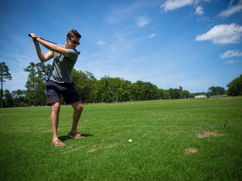 A young man in sunglasses is taking a golf swing on a sunny day at a driving range, with green grass and trees in the background.
