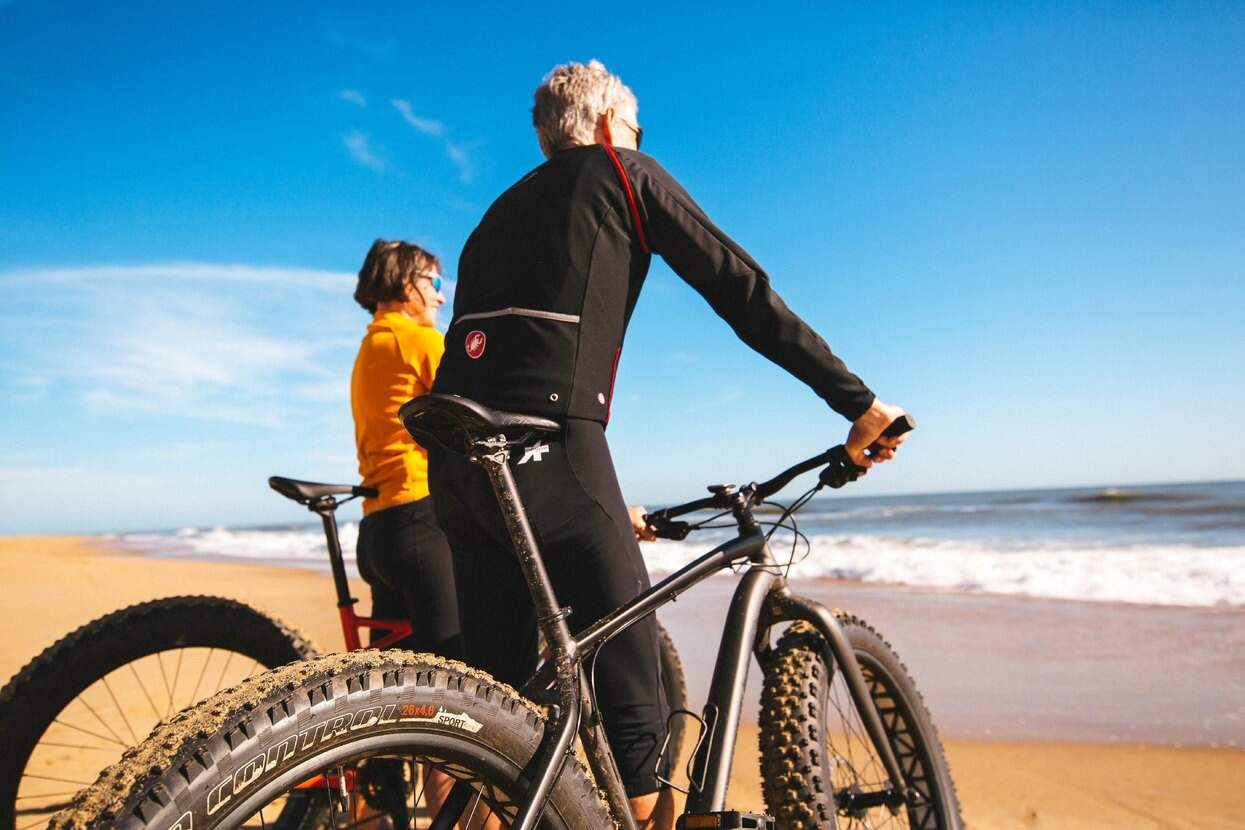 Two cyclists enjoying a beach view with their bikes, under a clear blue sky and near the ocean waves.
