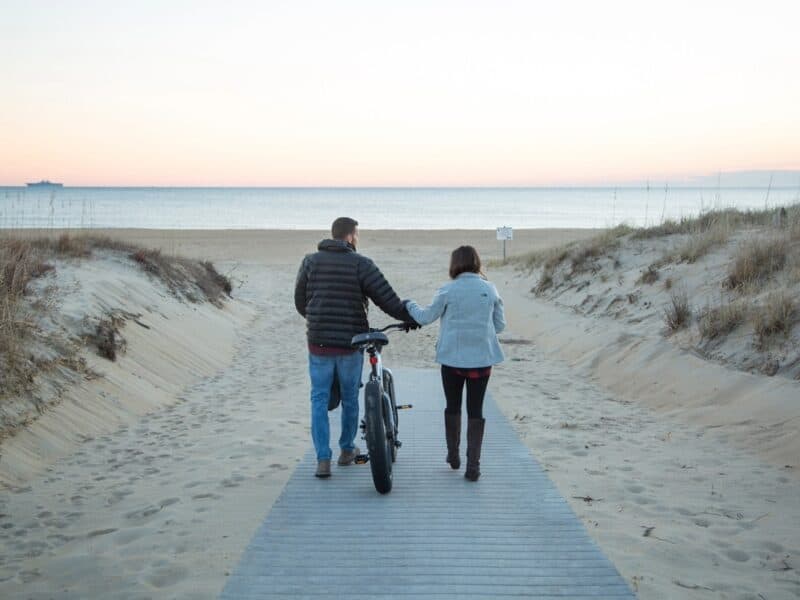 A couple walks hand-in-hand along a sandy path leading to the beach, with a bike beside them and a tranquil ocean view at sunset.