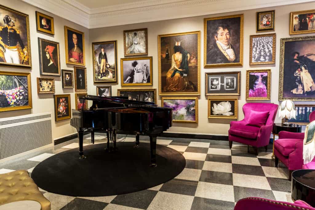 Elegant lounge featuring a grand piano surrounded by various framed artworks and plush pink chairs on a checkerboard floor.